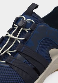 Αθλητικό παπούτσι σκούρου μπλε με πλέγμα στο πάνω μέρος, ελαστικά κορδόνια τύπου μπαγκάντα με κουμπί ρύθμισης και δύο φαρδιές υφασμάτινες λουρίδες πάνω από το πόδι.