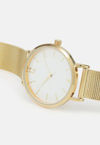 Montre avec bracelet en mailles dorées et boîtier rond. Le cadran blanc présente des index horaires fins. Inclut une couronne et une finition lisse. Design minimaliste.