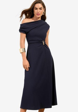 Kobieta w ciemnogranatowej sukience midi z odkrytymi ramionami, zapiętej złotą klamrą w talii, nosząca dużą złotą bransoletę na mankiet i złote kolczyki koła.