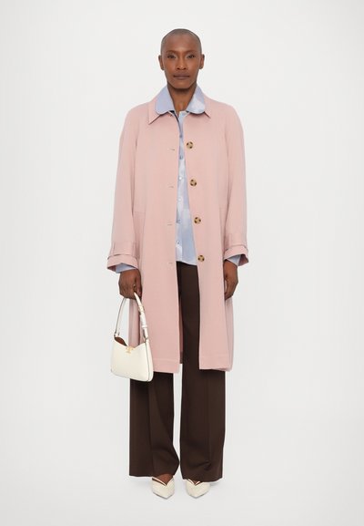 Lys pink lang frakke med store knapper, lagd over en lys blå skjorte. Bredde brune bukser og hvide hæle fuldender outfittet, med en hvid håndtaske.