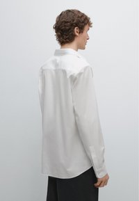 Witte blouse met lange mouwen, een gebogen zoom, klassieke kraag en manchetten met knopen, gemaakt van gladde stof, gezien van achteren.