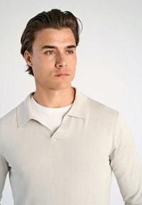 Jasno beżowa koszula polo z długim rękawem, wykonana z teksturowanej dzianiny, z dekoltem w kształcie litery V i płaskim kołnierzykiem na białej koszuli.