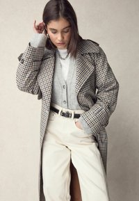 Trench coat a scacchi nei toni beige, marrone e navy. Indossato sopra un cardigan grigio e una maglietta bianca, abbinato a pantaloni beige a vita alta e una cintura nera.