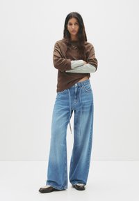 Weite blaue Denim-Jeans mit heller Waschung und Kordelzug. Kombiniert mit einem braunen Langarmshirt mit grauen Ärmelakzenten und einer dezenten Halskette.