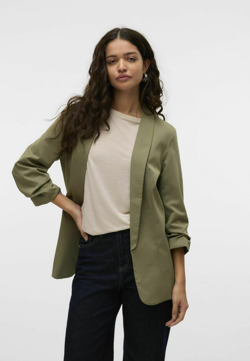 Olivgrön kavaj med en slät textur, öppen front och uppvikta ärmar, kombinerad med en ljusbeige topp och mörkblå jeans.