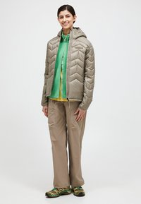 Lichtbruine pufferjack met chevronpatroon, groene fleece binnenlaag, beige broek en groene en zwarte schoenen. Al deze kleding is ontworpen voor buitengebruik.