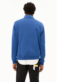 Sweatshirt en coton bleu avec un col haut, des poignets et un ourlet côtelés, et une coupe décontractée. Présente un look superposé avec une chemise blanche en dessous.
