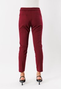 Pantaloni dark red slim-fit con tasche posteriori, indossati con sandali neri con tacco, mostrati da dietro su uno sfondo bianco.