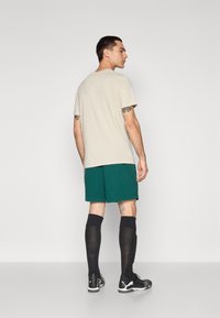 Beżowa koszulka z krótkim rękawem, zielone szorty, czarne skarpety do kolan, czarne sneakersy. Minimalistyczny design, regularny krój, widoczne tatuaże na ramionach.