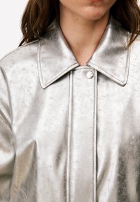 Veste argentée métallique à texture lisse, grand col, fermeture à pression et détails de coutures visibles. Vue rapprochée de l'avant.