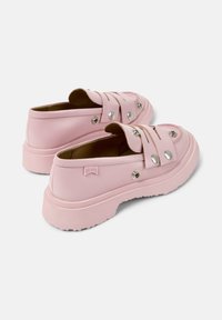 Rosa slip-on skor med en kraftig sula, prydda med dekorativa remmar och metallnitar, presenterade mot en enfärgad vit bakgrund.