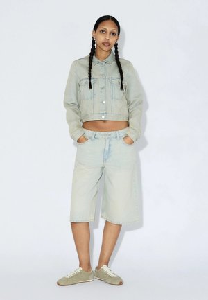 Kvinde med langt flettet hår iført lys denimjakke, matchende shorts til knæet og beige sneakers, står med hænderne i lommerne.