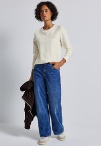 Krémová mikina s nápisem "ATELIER", kombinovaná s širokými modrými džíny. Model drží hnědou bundu. Koutfit doplňují tenisky.