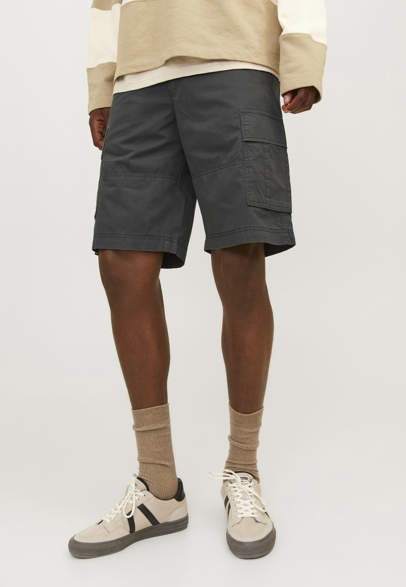 Uomo che indossa pantaloni cargo grigi scuri, camicia beige a maniche lunghe, calze beige e scarpe da ginnastica beige con strisce nere su uno sfondo neutro.