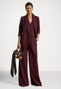 Μπορντό κοστούμι τριών τεμαχίων με σακάκι ραμμένο στα μέτρα, γιλέκο και παντελόνια με φαρδιά μπατζάκια. Το μοντέλο κρατά μια τσάντα με καφέ και μοτίβο, συνοδευόμενη από ένα κασκόλ.