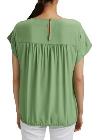 Femme portant un chemisier vert à manches courtes, ample, avec une ouverture en forme de trou à bouton au dos et un tissu froncé au niveau du yoke et de l'ourlet, associé à un pantalon blanc.