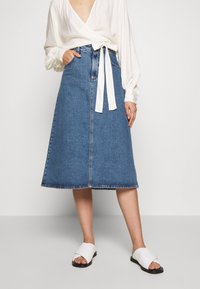 Denimová áčková sukně ve světle modré barvě, se zapínáním na knoflíky vpředu a bočními kapsami. Nosit s bílým překládaným topem a bílými sandály.