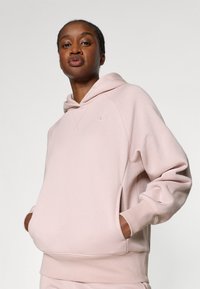 Sweatshirt à capuche rose clair avec une coupe décontractée, des poignets côtelés et une poche avant. Présente un logo subtil sur la poitrine et une texture douce.