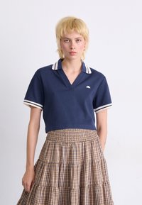 Navy cropped polo shirt with a V-neck, featuring white striped cuffs and a small logo. Paired with a beige plaid tiered skirt.