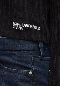 Czarny sweter z prążkowanego materiału z białym logo "KARL LAGERFELD JEANS"; zestawiony z ciemnoniebieskimi dżinsami, które mają srebrny guzik na kieszeni.