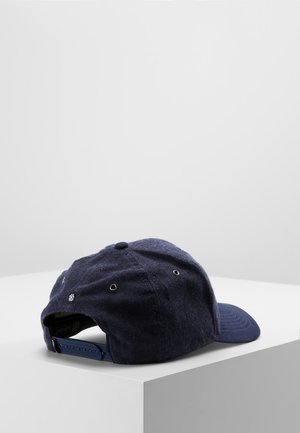 Casquette de baseball bleu foncé avec une sangle plastique réglable reposant sur une surface blanche sur un fond gris clair uni.