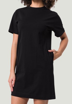 Vrouw draagt een zwarte T-shirtjurk met korte mouwen en knielengte, met één hand in een zijzak, kijkend naar voren tegen een effen achtergrond.
