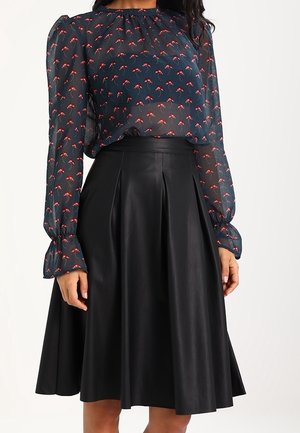 Frau trägt eine transparente marineblaue Bluse mit rotem Blumenmuster und langen, aufgepufften Ärmeln, kombiniert mit einem hoch taillierten schwarzen, plissierten Midirock.
