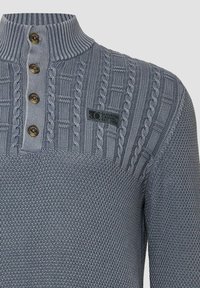 Hellgrauer gestrickter Pullover mit hohem Kragen, der horizontale und vertikale Muster aufweist, vier braune Knöpfe und ein schwarzes Logopatch hat.