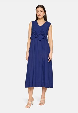 Vrouw draagt een mouwloze marineblauwe midi-jurk met bloemige kanten details op het lijfje, gecombineerd met beige sandalen met hak, staand tegen een effen achtergrond.