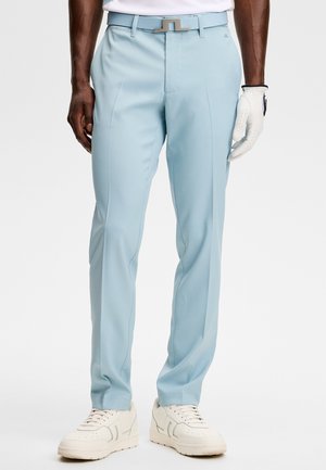 Homme portant un pantalon ajusté bleu clair, ceinture blanche, baskets blanches et gant de golf blanc à la main droite.