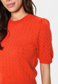 Stickad tröja i klar orange, med ett texturerat zigzag-mönster och puffärmar i kort längd. Mjuk, lättviktsmaterial.