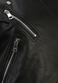 Giacca in pelle nera con cerniere e accessori in argento. Presenta un materiale strutturato e molteplici tasche con cerniere. Vista ravvicinata.