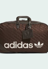 Brown duffel bag with repeating "adidas" pattern, textured fabric, silver logo, sturdy handles, and a small attached tag featuring the logo.