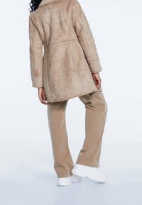 Beige nepbontjas met een brede kraag, voorzien van stiksels. Gepaard met ruime beige broek en witte chunky sneakers.