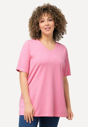 Vrouw met krullend haar, gekleed in een roze T-shirt met korte mouwen en een V-hals, en blauwe jeans, staand tegen een effen lichte achtergrond, vriendelijk glimlachend.