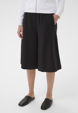 Person trägt schwarze Culotte-Shorts mit weitem Bein, schwarze Slipper und eine weiße Reißverschlussjacke mit Händen in den Taschen.