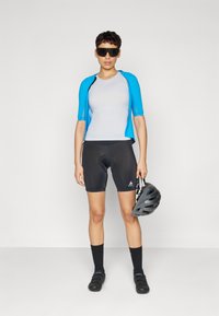 Radbekleidung bestehend aus einem blauen kurzärmligen Trikot, einem grauen enganliegenden Oberteil, schwarzen Radhosen, schwarzen Socken und einem grauen Helm, der in der Hand gehalten wird.