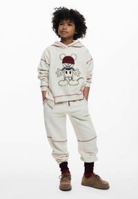Sudadera color crema con costuras rojas que presenta un personaje de dibujos animados; combinada con unos joggers a juego. Conjunto hecho de un tejido suave. Zapatos marrones.