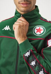Chaqueta deportiva verde con cuello alto, acentos rojos, cierre de cremallera y parche de logo con una estrella roja. Puños acanalados y mangas con estampado.