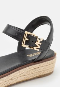 Černé klínové sandály s nastavovacími popruhy se zlatou sponou a pletenou juteovou podrážkou, které se vyznačují elegantním a moderním designem.