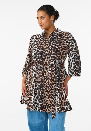 Femme aux cheveux bouclés portant une tunique ceinturée à imprimé léopard sur un jean bleu, regardant sur le côté sur fond blanc.