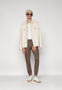 Beige overhemdjas met twee borstzakken, wit T-shirt, bruine cargobroek en witte sneakers met bruine zolen. Accessoires zijn onder andere een zonnebril en een beige beanie.