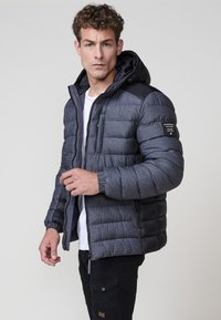Padded jakna u tamno sivoj boji s kapuljačom, koja sadrži kombinaciju teksturiranog i glatkog materijala, zatvaranje na patentni zatvarač i oznaku s logotipom na rukavu.