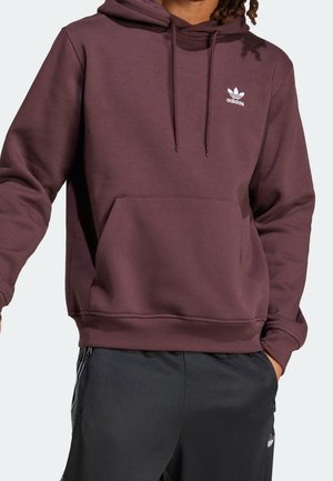 Personne portant un sweat à capuche Adidas bordeaux avec un logo blanc et un pantalon Adidas noir avec des rayures blanches et un logo, debout avec une main dans la poche.