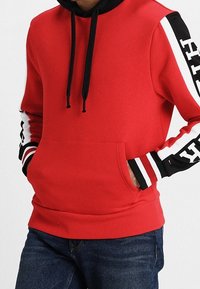 Rode hoodie met zwarte koorden en witte, zwarte en rode gestreepte mouwen, voorzien van vetgedrukte zwarte tekst op de armen, gedragen met blauwe jeans.