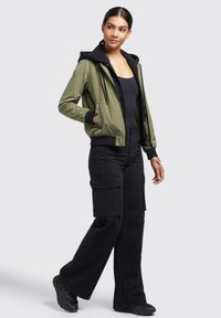 Jachetă bomber de culoare verde măslinie cu glugă și mansete negre, având închidere tip fermoar și două buzunare laterale, purtată împreună cu pantaloni cargo negri și adidași negri.