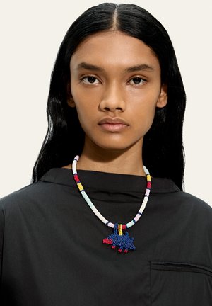 Młoda osoba z długimi czarnymi włosami ubrana w czarny top i wielokolorowy naszyjnik z koralików z niebiesko-czerwonym wisiorkiem w kształcie zwierzęcia.