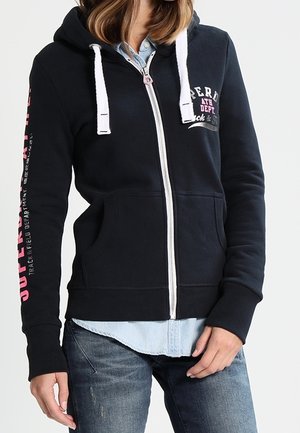 Femme portant un sweat à capuche noir zippé avec des cordons blancs, des lettres roses et blanches, superposé à une chemise bleu clair boutonnée et un jean bleu.