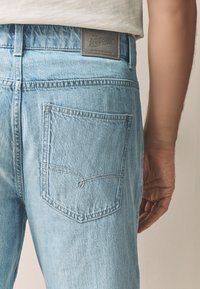 Jeans in denim azzurro chiaro con una tasca posteriore decorata da un motivo cucito. Etichetta grigia visibile in vita, con dettagli del marchio.