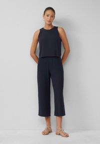 Navy jumpsuit met mouwloos ontwerp, ontspannen pasvorm, cropped brede benen, lichtgewicht stof en subtiele glans. Gepaard met platte sandalen.